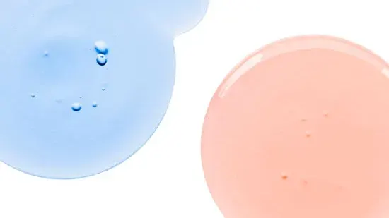 Nærbilde av to dråper væske på en hvit bakgrunn, en blå og en rosa.