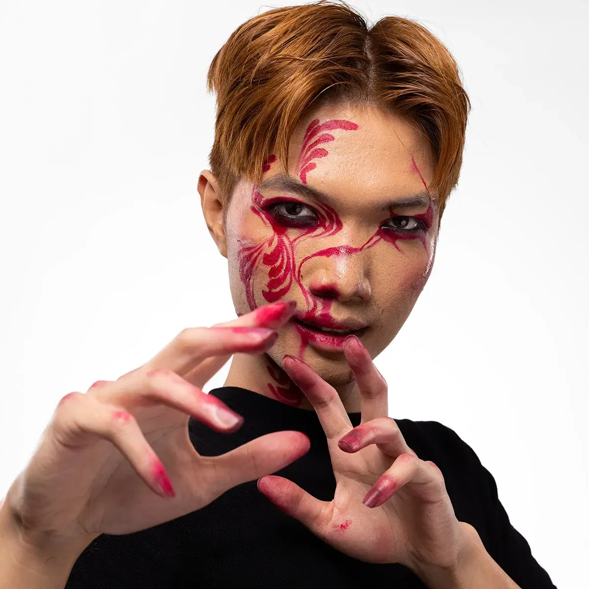En person med dramatisk röd ansiktsmålning i svepande linjer över ögon, kinder och mun, kombinerat med intensiv ögonmakeup och rödfärgade fingertoppar som ger en konstnärlig, effektfull look.
