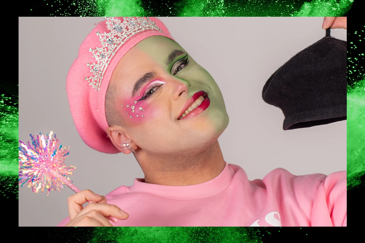 Memo med rosa smink och glitter i rosa klädsel, leendes in i kameran, mot en grå bakgrund med grön ram.