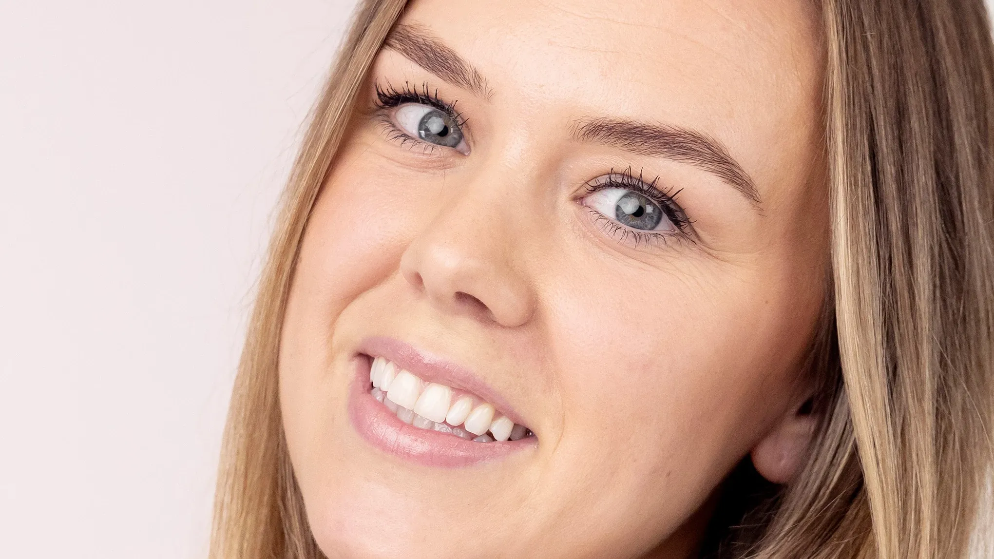 En kvinne med sminke som smiler inn i kameraet, mot en rosa bakgrunn.