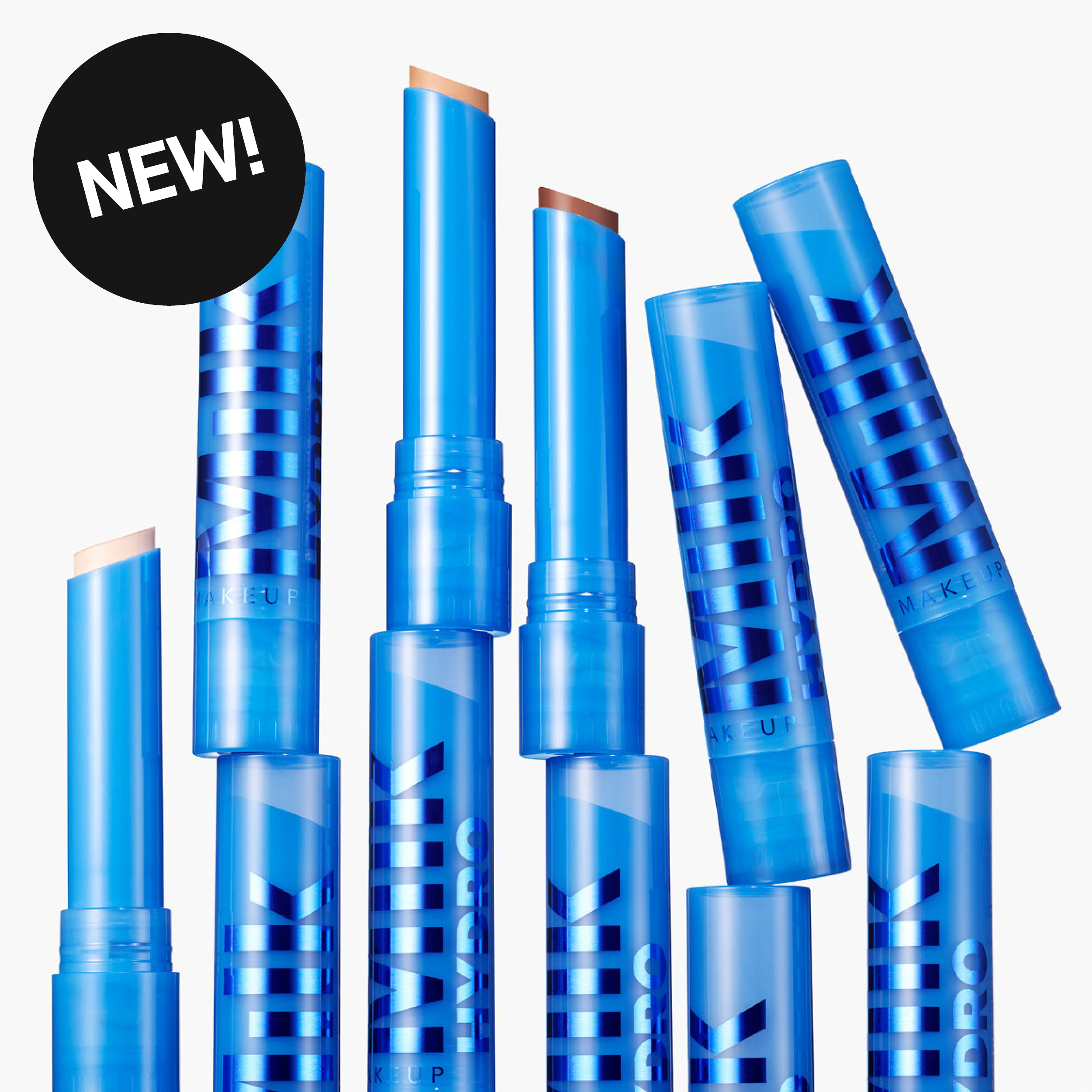 Milk Makeup -meikkituotteet sinisissä pakkauksissa tekstillä “MILK MAKEUP HYDRO”. Useita stick-muotoisia tuotteita näkyy avattuina vaaleaa taustaa vasten sekä pyöreä merkki tekstillä “NEW!”.