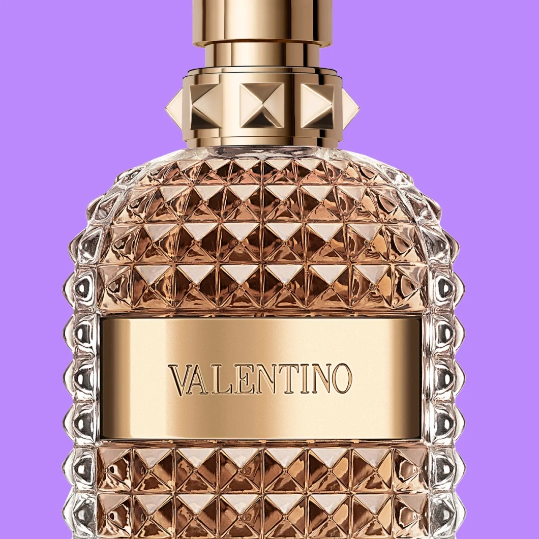 Lähikuva Valentino-hajuvedestä vaalean roosanvärisessä lasipullossa, jossa on pyramidinmuotoisia niittejä, kullanvärisiä yksityiskohtia ja näkyvä teksti ”VALENTINO”, violetilla taustalla.