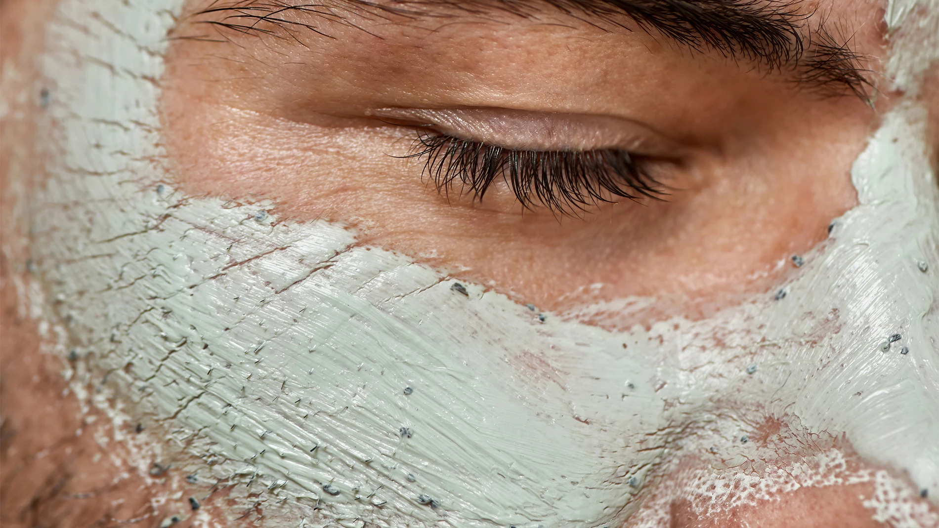 En persons ansikte med slutna ögon som visar en ansiktsmask av grön lera som applicerats på pannan, näsan och kinderna.