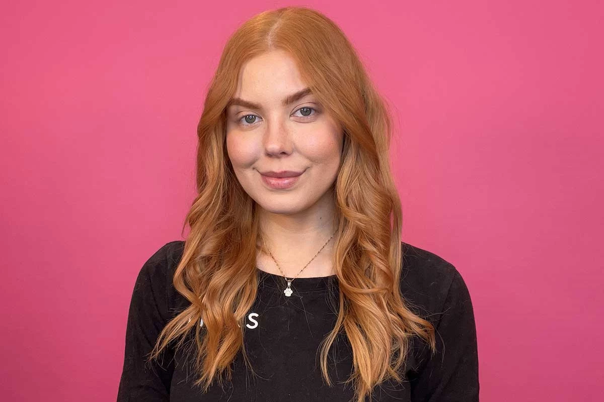 En person med långt, rött hår står framför en rosa bakgrund och har på sig en svart tröja med texten "KICKS". Håret är stylat i lösa lockar.