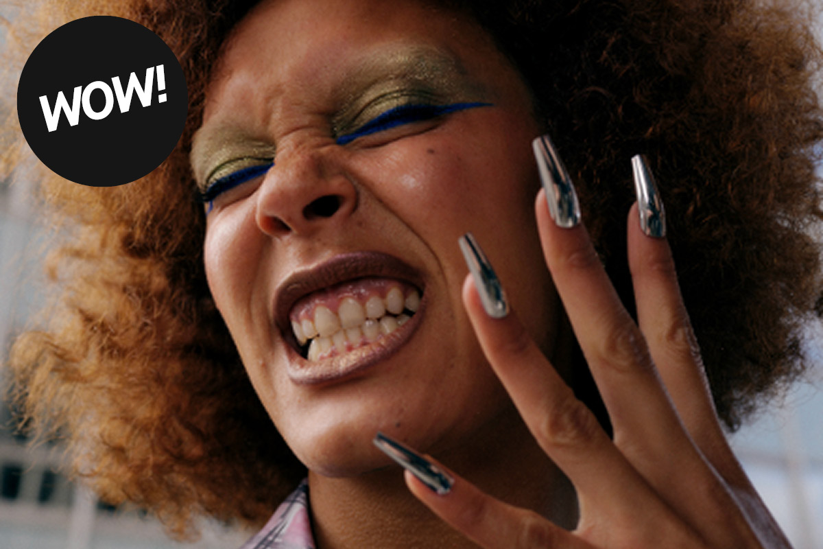 Kuva henkilöstä, jolla on kiharat hiukset ja musta ympyrä, jossa lukee "WOW!" vasemmassa yläkulmassa. Henkilöllä on pitkät, metalliset kynnet ja hän pitää kättä ylhäällä.