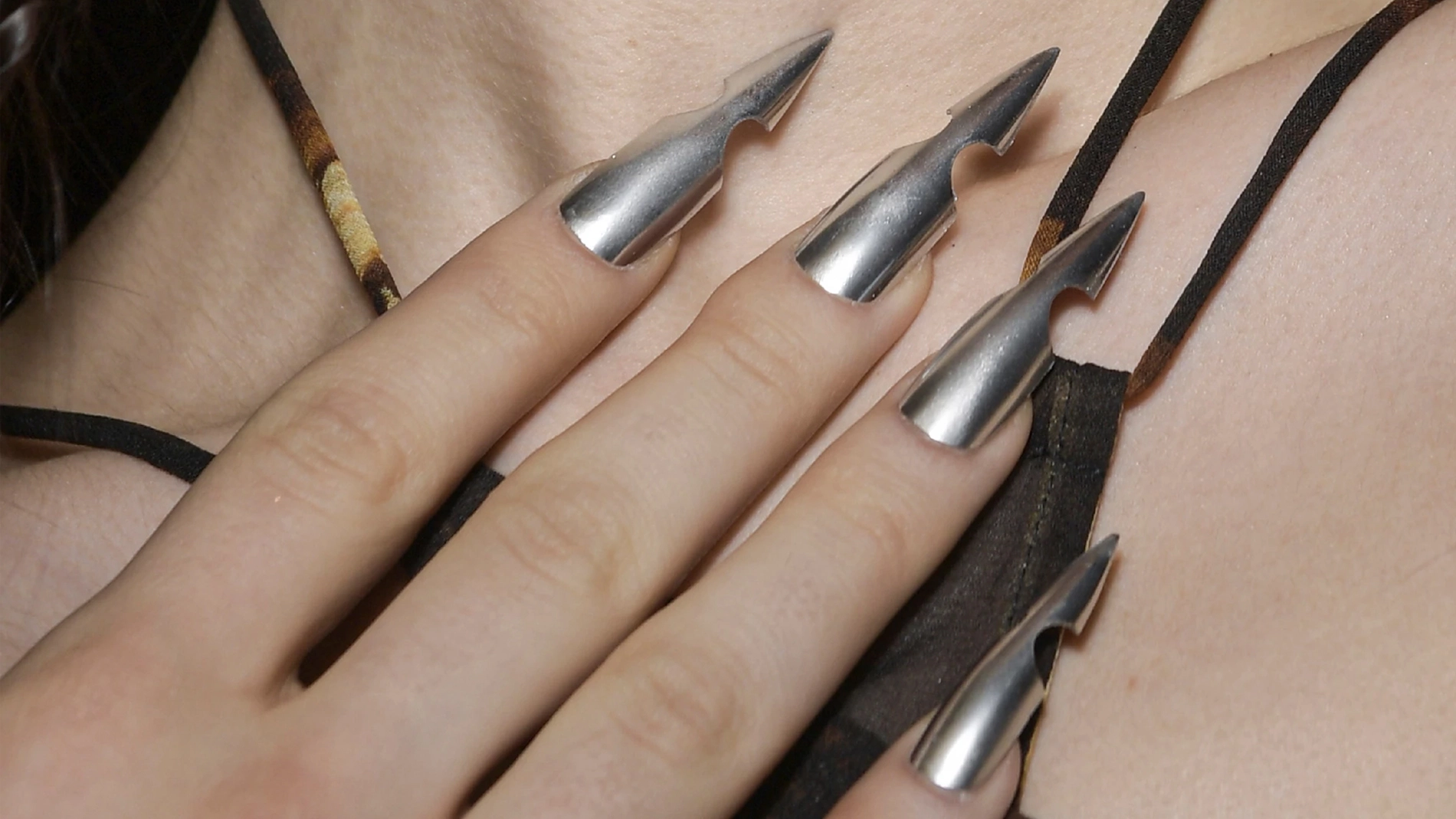 Närbild på en hand med långa, metalliska naglar med unika, spetsiga och urholkade design. Naglarna har en glänsande silverfärg.