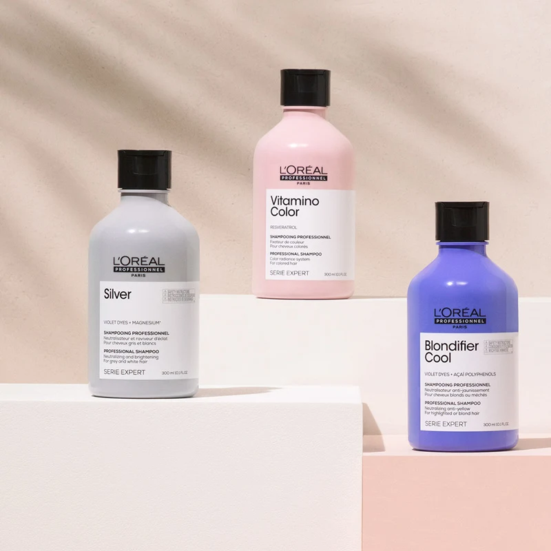 Tre schampon från L'Oréal Professionnels Serie Expert står uppradade på vita kuber mot en beige bakgrund. En flaska är grå, en är rosa och en är lila. Alla flaskor har svarta lock och vita etiketter med svart text.
