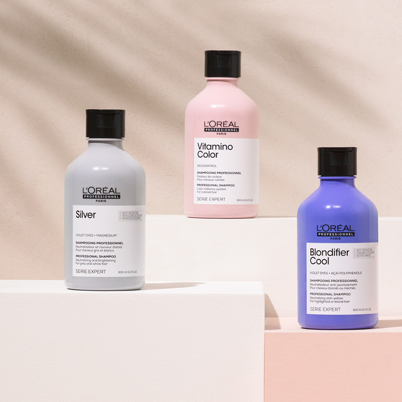 Kolme L'Oréal Professionnelin Serie Expert -sarjan shampoota on rivissä valkoisilla kuutioilla beigeä taustaa vasten. Yksi pullo on harmaa, yksi vaaleanpunainen ja yksi violetti. Kaikissa pulloissa on mustat korkit ja valkoiset etiketit, joissa on musta teksti.