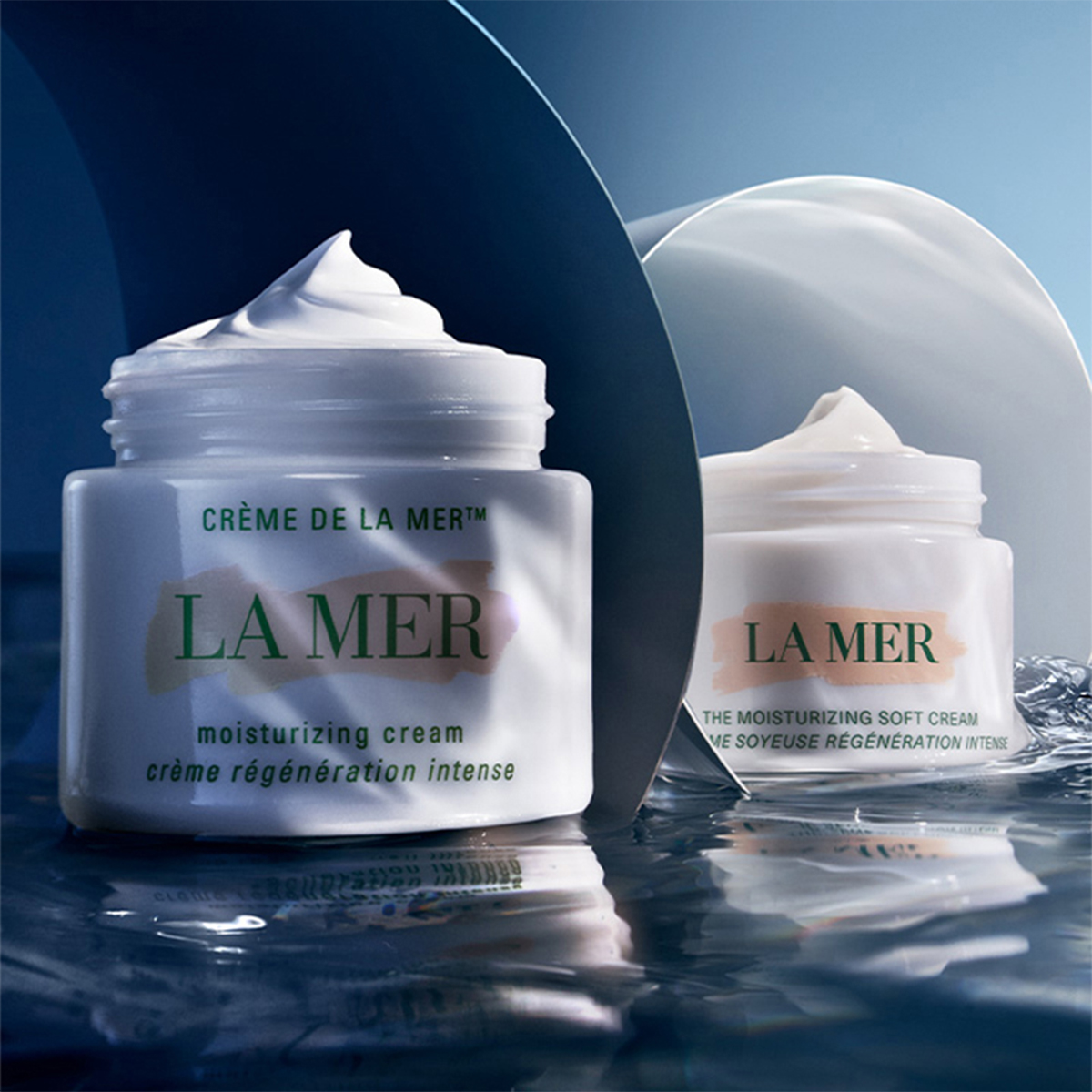 To krukker med fuktighetskremer fra La Mer plassert på en reflekterende flate med blå bakgrunn som fremhever produktenes luksuriøse og fuktighetsgivende egenskaper.