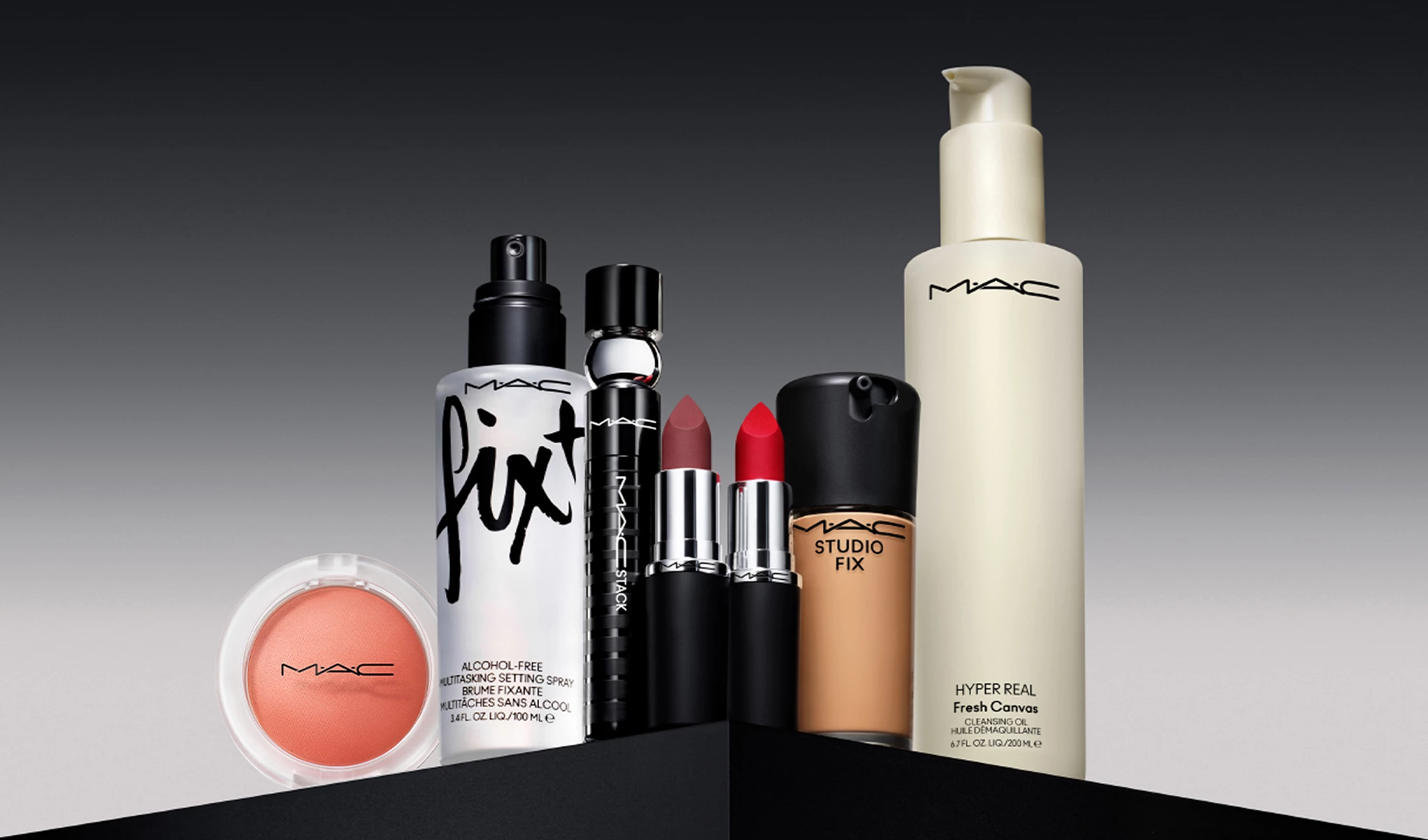 MAC Cosmetics -meikkituotetta on rivissä tummanharmaalla taustalla. Tuotteilla on erilaiset mustat, valkoiset ja beiget pakkaukset, joissa MAC-logo on selvästi näkyvissä.