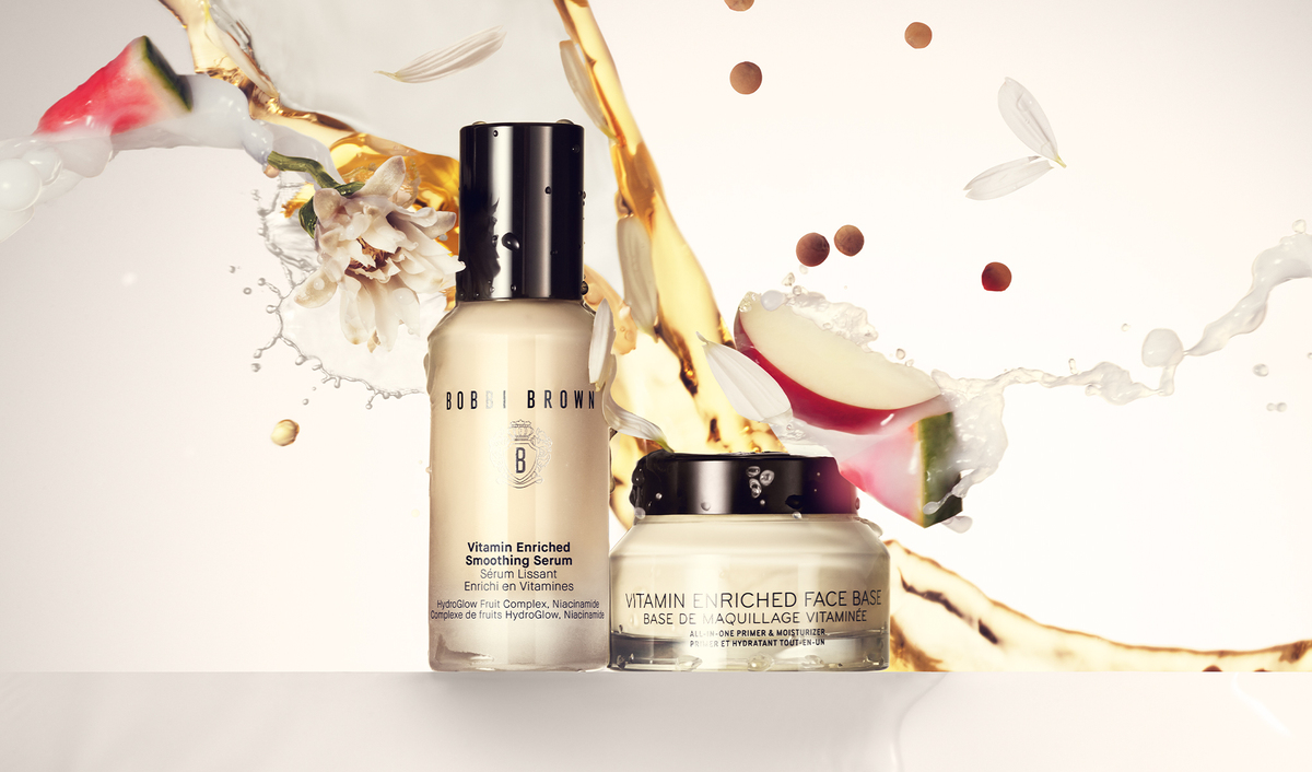 Bobbi Brownin Vitamin Enriched Smoothing Serum ja Vitamin Enriched Face Base. Seerumi on beigessä pullossa, jossa on musta kansi, ja kasvovoide beigessä purkissa, jossa on musta kansi. Pohja on täytetty roiskeilla nestettä, kukan terälehdillä ja hedelmänpaloilla, jotka luovat dynaamisen ja värikkään vaikutelman.