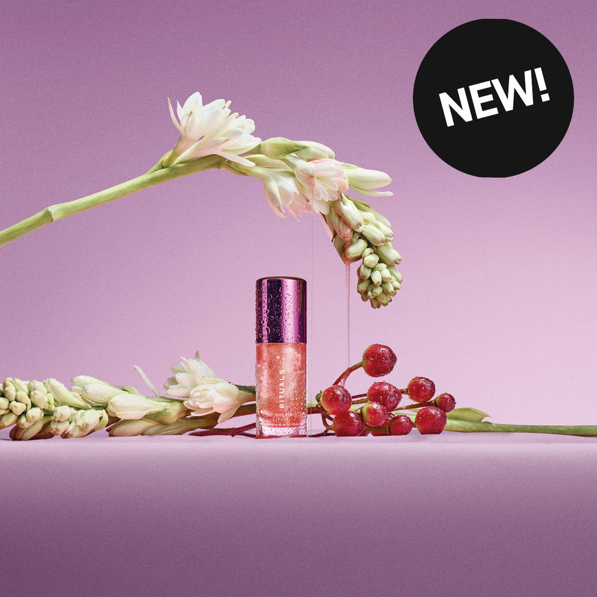 Rituals leppeprodukt i en liten flaske med skimrende rosa innhold og teksten “RITUALS”, plassert blant blomster og røde bær mot rosa bakgrunn. En svart sirkel med teksten “NEW!” vises i bildet.