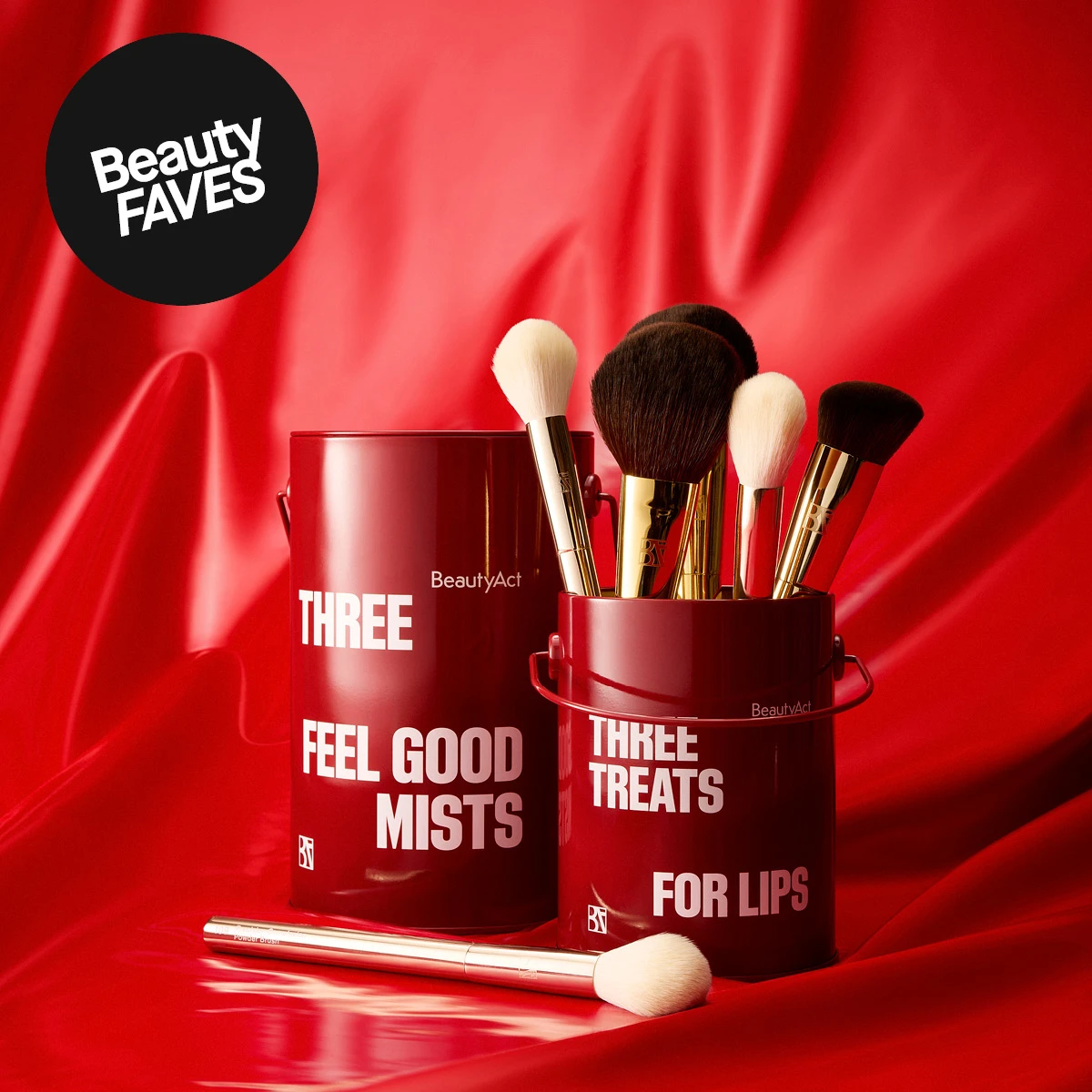 Kaksi punaista BeautyAct-säiliötä: toisessa 'Three Feel Good Mists', toisessa 'Three Treats for Lips' ja meikkisiveltimiä. Yksi sivellin edessä. Ylhäällä vasemmalla: 'Beauty FAVES'.
