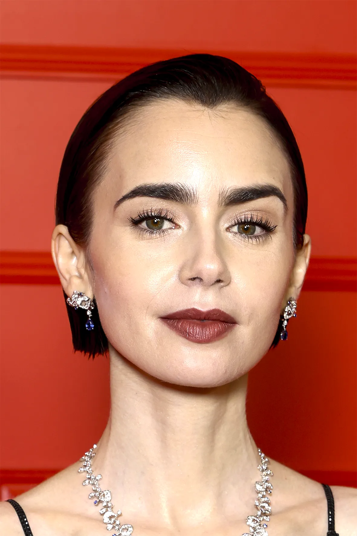 Nærbilde av person med oppsatt mørkt hår og naturlig makeup med jevn hud, markerte bryn, mascara og brunrosa leppestift. Personen har glitrende øredobber og et matchende smykke mot en rød bakgrunn.