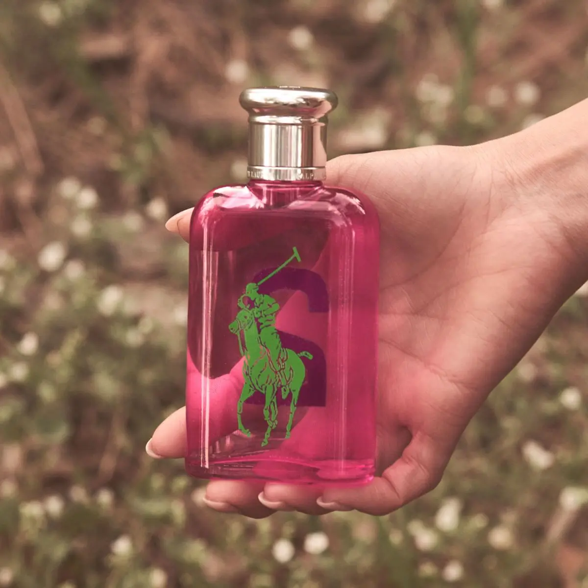 En hand håller en rosa Ralph Lauren parfymflaska utomhus med natur i bakgrunden.