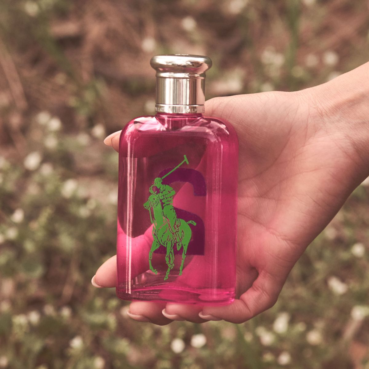 En hånd holder en rosa Ralph Lauren parfymeflaske utendørs med natur i bakgrunnen.