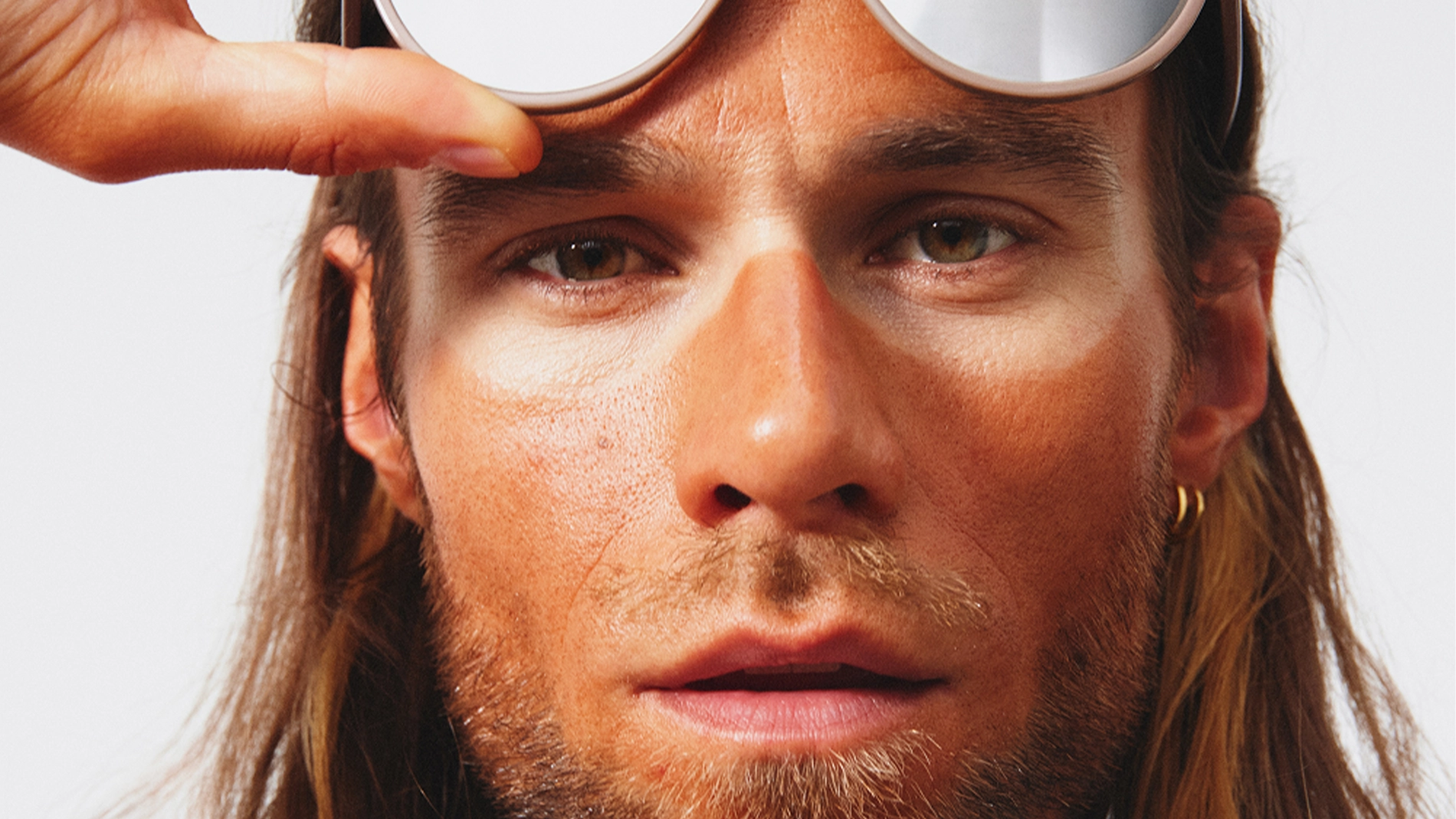 Nærbilde av en person som løfter opp et par store solbriller med speilglass og beige innfatning. Ansiktet er dekket av solbrun foundation eller bronzer, med tydelig lysere hud rundt øynene der solbrillene har sittet.