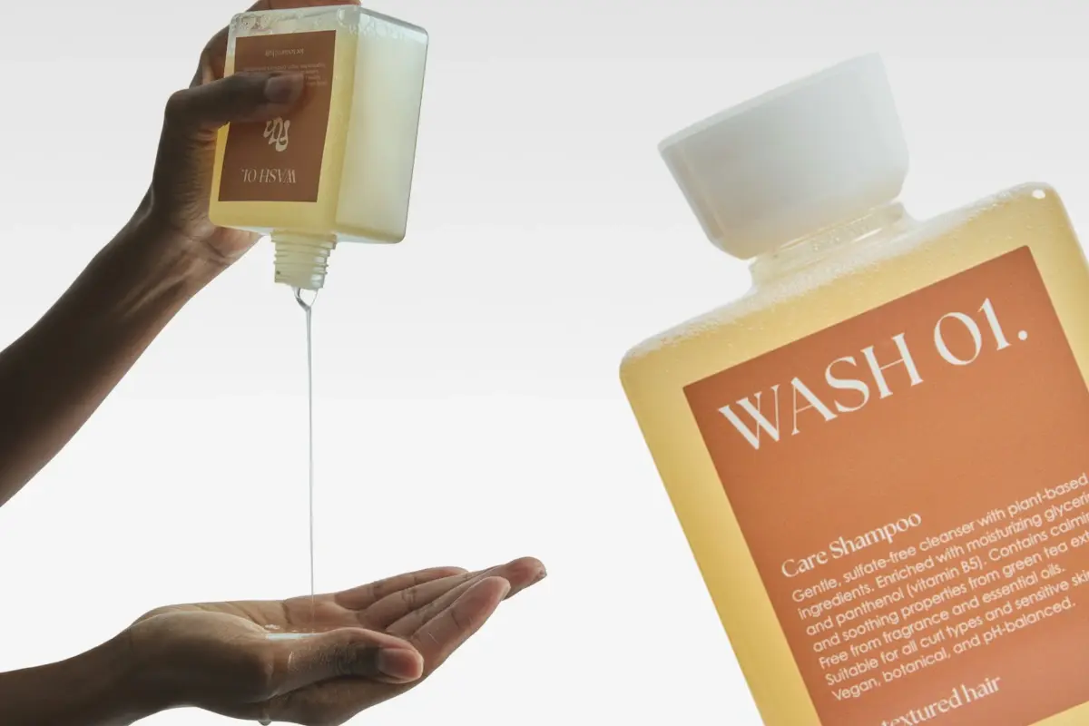 En fyrkantig flaska med WASH 01. Care Shampoo, en skonsam rengöring, häller vätska i en öppen hand.