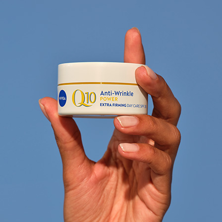 En hand som håller en burk från Nivea mot en klar blå himmel. Burken är vit med blå och gul text som visar produktnamnet. Handen är elegant positionerad och framhäver produkten mot den enkla bakgrunden.