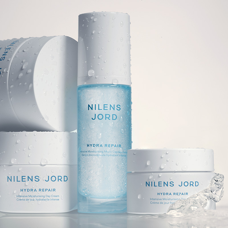 Nilens Jord Hydra Repair -tuotteet, jotka sisältävät intensiivisesti kosteuttavaa päivävoidetta ja mikrokapseleita sisältävää seerumia. Tuotteet ovat valkoisissa ja sinisissä pakkauksissa, ja niissä on vesipisaroita.
