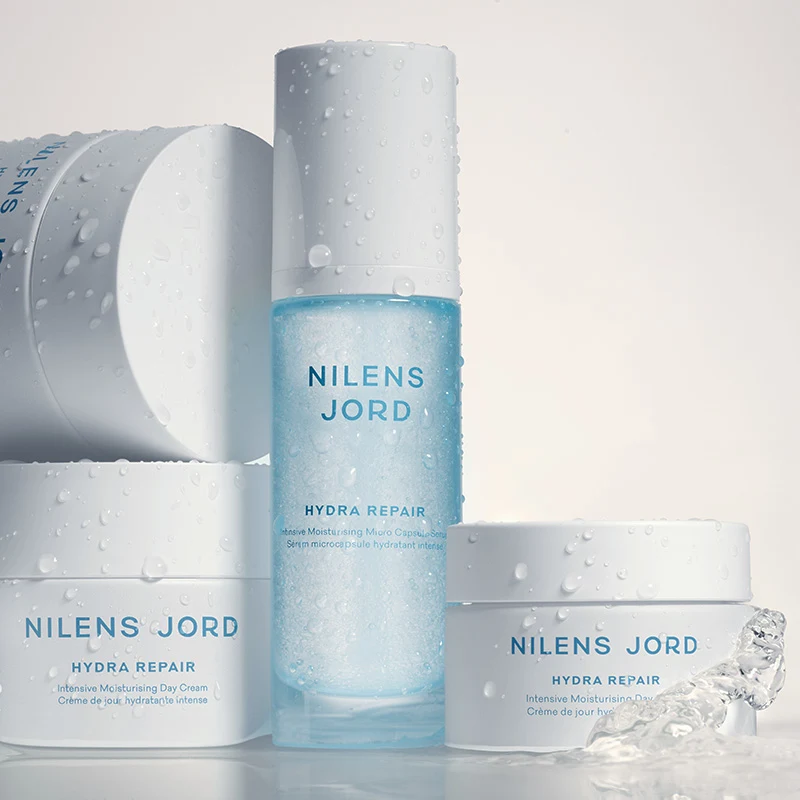 Nilens Jord Hydra Repair -tuotteet, jotka sisältävät intensiivisesti kosteuttavaa päivävoidetta ja mikrokapseleita sisältävää seerumia. Tuotteet ovat valkoisissa ja sinisissä pakkauksissa, ja niissä on vesipisaroita.