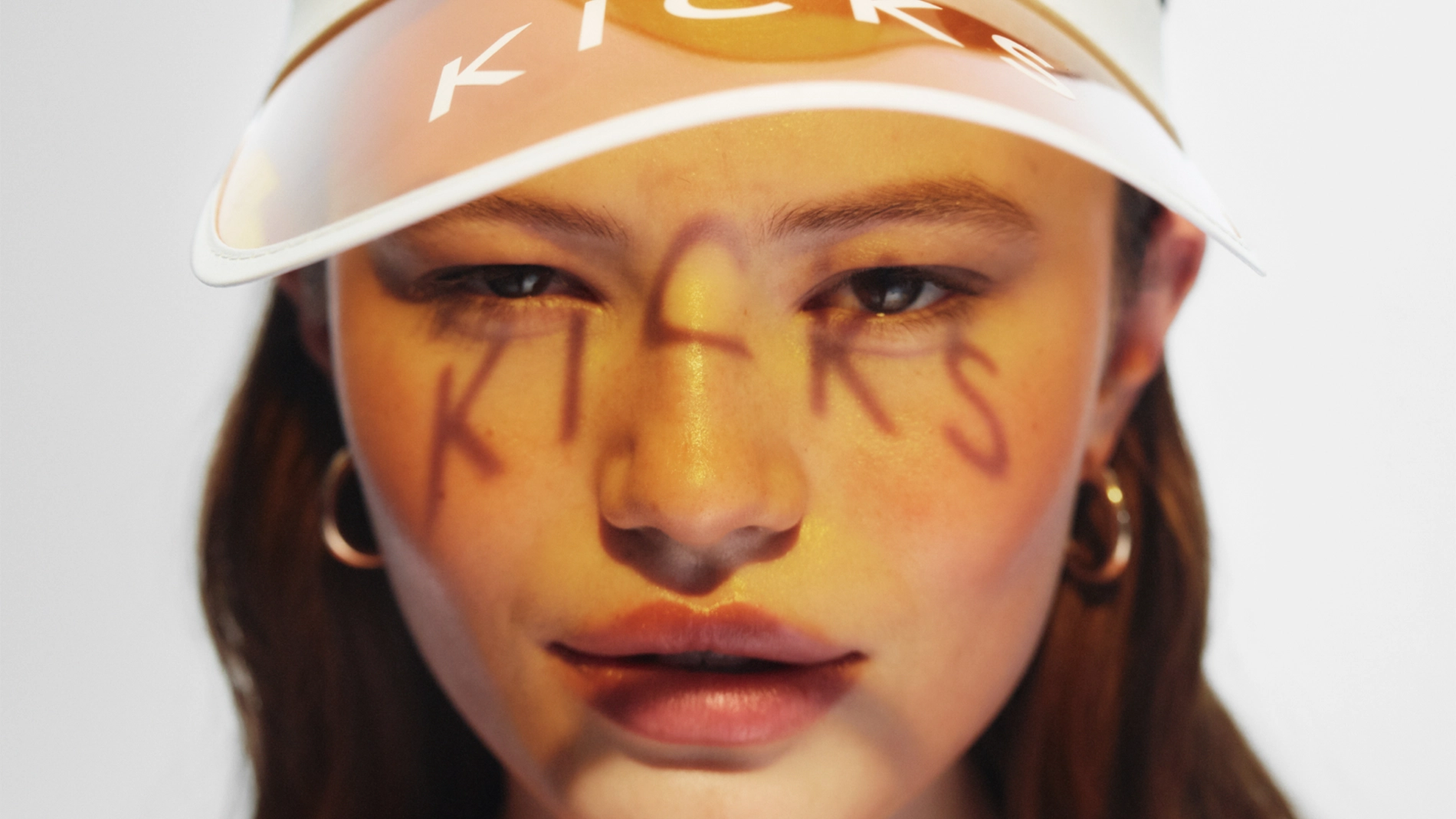 Nærbilde av ansikt med kreativ sminke i gule og oransje toner og glansfulle lepper, der ordet “KICKS” er malt over kinn og nese. Personen har på en transparent solskjerm med teksten “KICKS” og store hoop-øredobber.