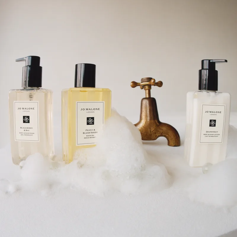 Tre flaskor med Jo Malone-produkter placerade på ett skummigt underlag framför en gammaldags kran. 