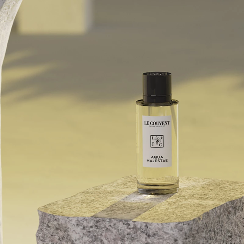 En klar glasflaska med parfym märkt "Le Couvent Aqua Majestae" står på en ljus stenblock i ett beige rum.