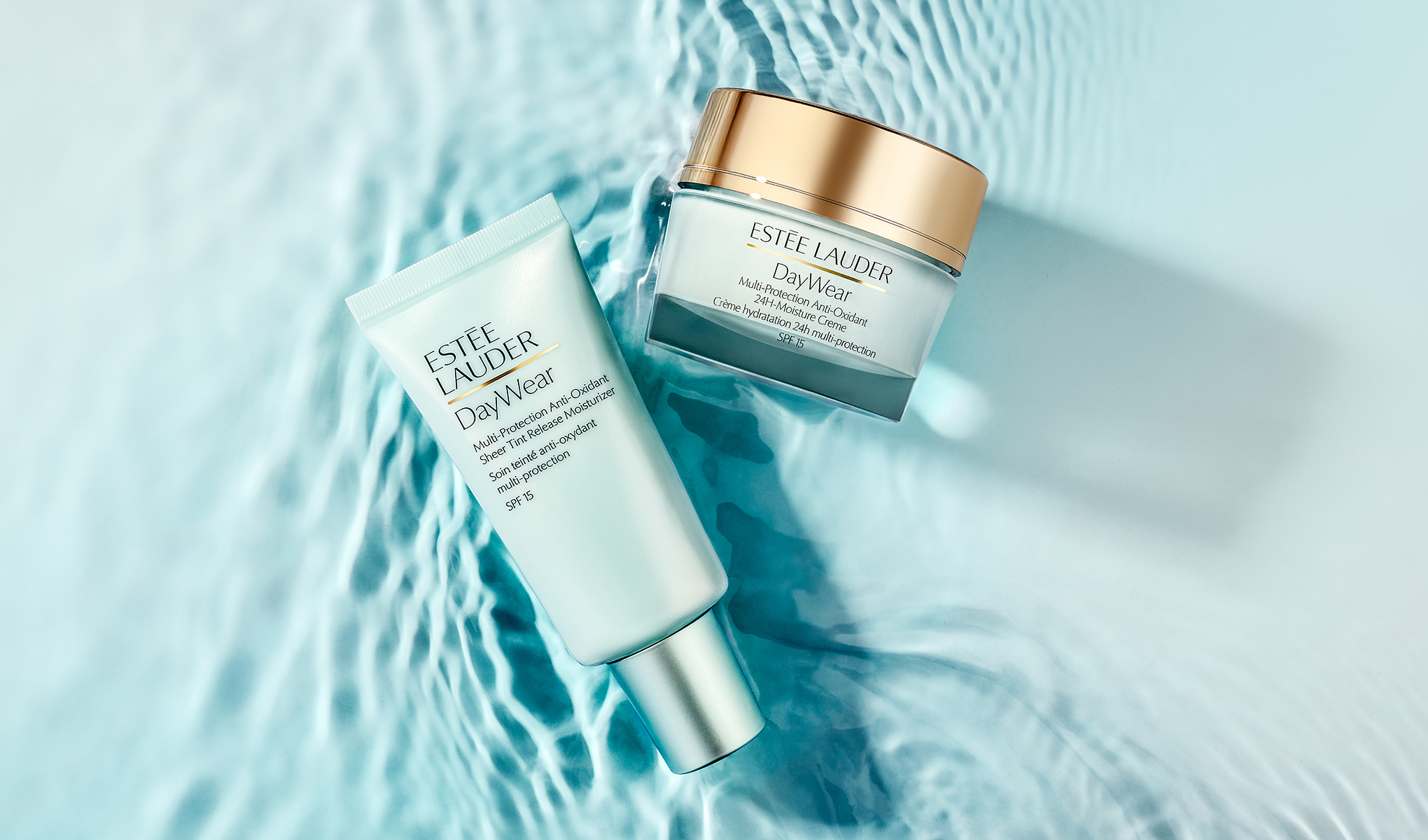To hudpleieprodukter fra Estée Lauder DayWear ligger på en lyseblå vannflate med bølgende refleksjoner. En tube med teksten «ESTÉE LAUDER DayWear Multi-Protection Anti-Oxidant Sheer Tint Release Moisturizer SPF 15» vises ved siden av en krukke merket «ESTÉE LAUDER DayWear Multi-Protection Anti-Oxidant 24H-Moisture Creme SPF 15».
