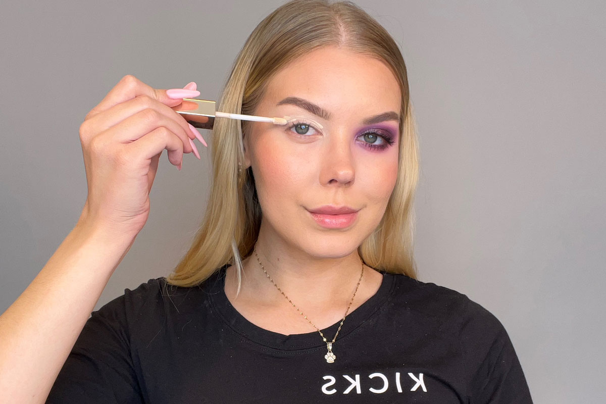 Kuva naisesta, jolla on violetti luomiväri toisessa silmässään, ja hän levittää luomivärin pohjamaalia toiselle silmäluomelleen.