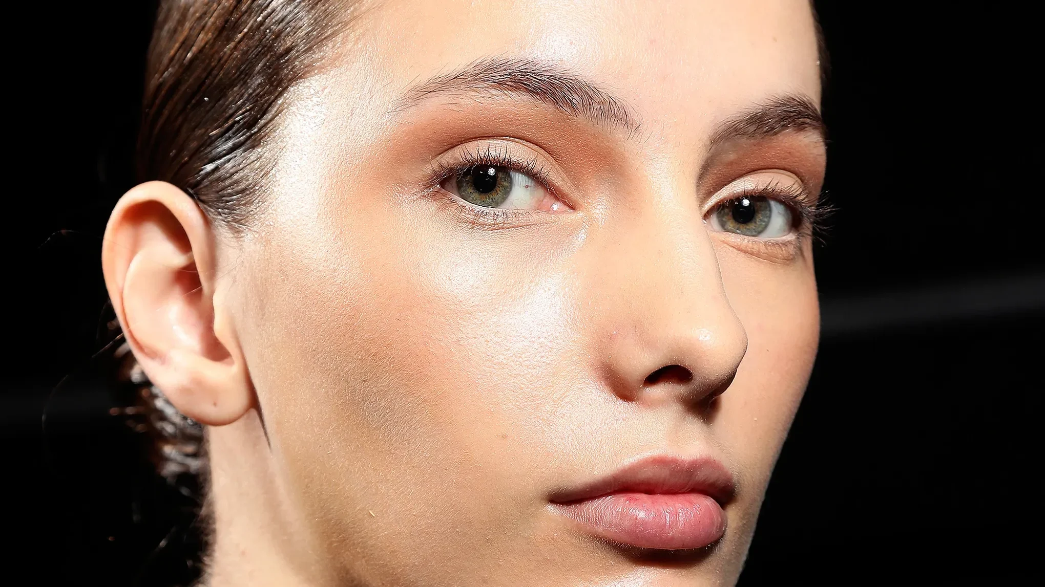 Närbild på en kvinna med återfuktad hud, naturlig makeup och glowig finish mot mörk bakgrund.