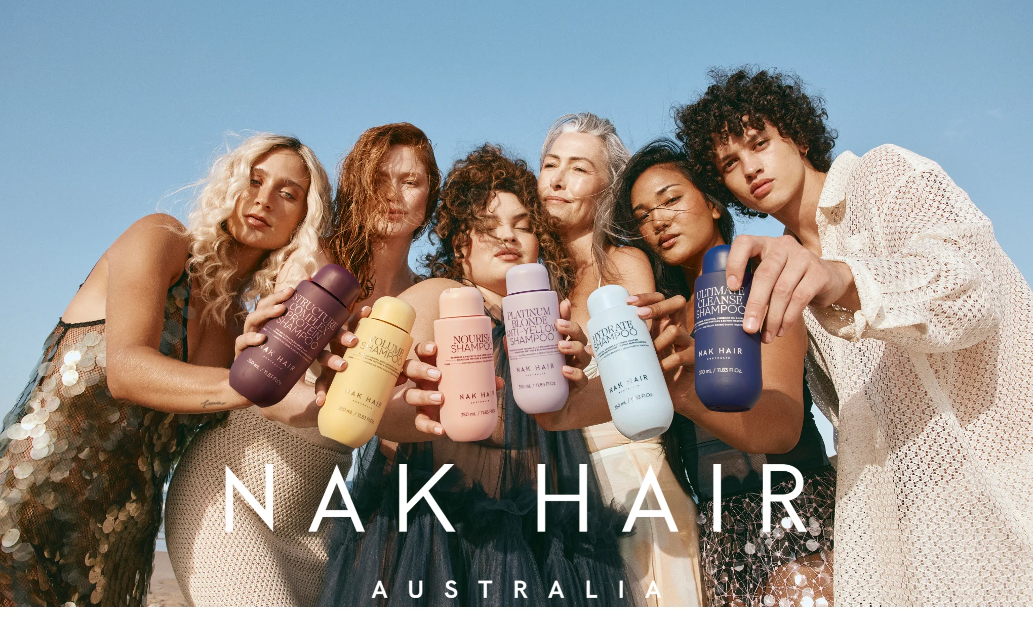 Ryhmä ihmisiä pitelee värikkäitä NAK Hair -shampoopulloja ulkona.