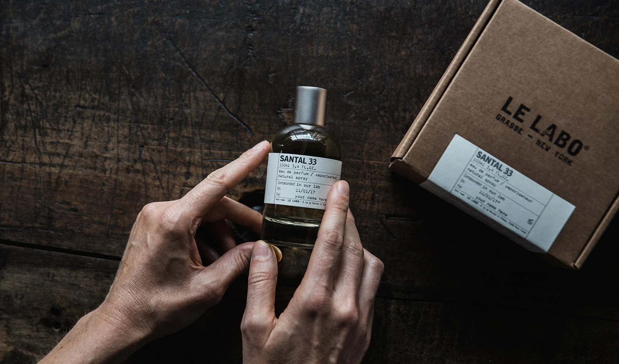 En person håller i en flaska med parfymen Santal 33 från Le Labo. Flaskan har en vit etikett med text och står på ett mörkt träbord. Till höger om flaskan finns en brun kartong med Le Labo-logotypen och en etikett som matchar flaskans.