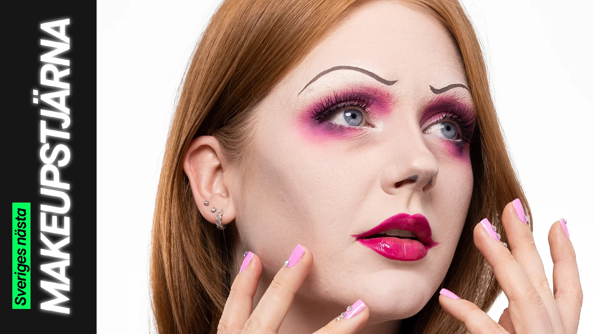 En person med en kreativ makeuplook i rosa toner, med markerad ögonmakeup, långa fransar, skarpt målade tunna ögonbryn och glansiga ceriserosa läppar, där händerna med rosa naglar ramas in omkring ansiktet.