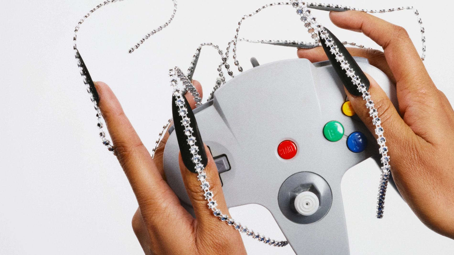 Två händer håller i en grå spelkontroll med färgade knappar. Över händerna och kontrollen ligger silverfärgade kedjor och glittrande dekorationer mot en ljus bakgrund.