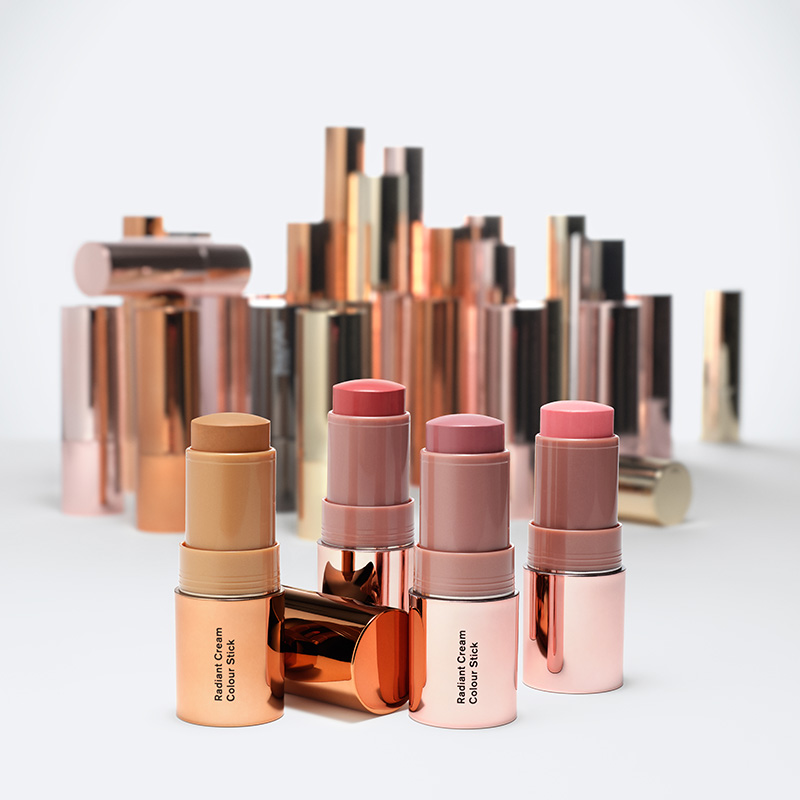Radiant Cream Colour Stick BeautyAct by KICKS -väritikkuja eri sävyissä, kuten beige, vaaleanpunainen ja ruskea. Pakkauksessa on metallinvärisiä värejä, kuten kultaa, kuparia ja vaaleanpunaista. Taustalla on valkoista ja taustalla on lisää pakkauksia, mikä luo tyylikkään ja ylellisen tunnelman.
