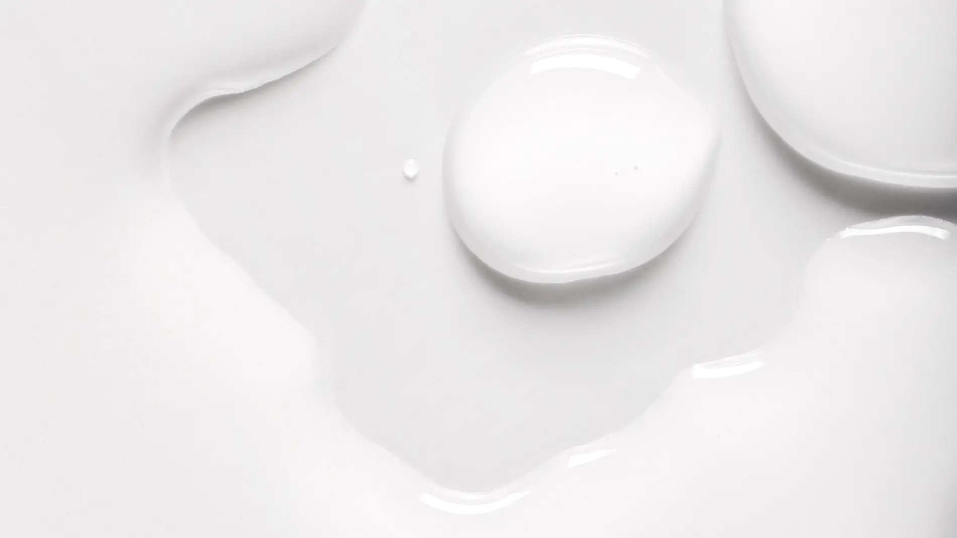 Nærbilde av hvit hudpleie med dråper og myk, glansfull serum- eller lotiontekstur mot hvit bakgrunn. Den kremede konsistensen fremhever et fuktighetsgivende skincare-produkt med lett og silkemyk finish.