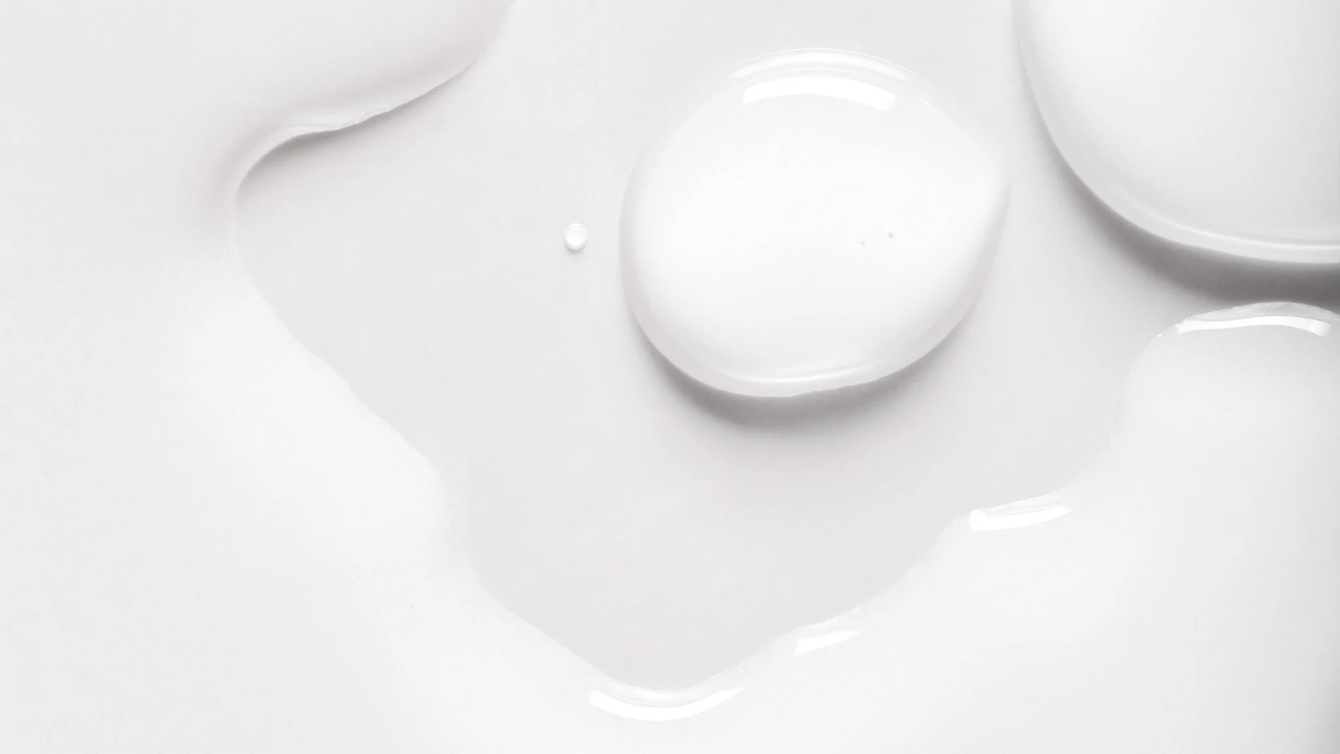 Nærbilde av hvit hudpleie med dråper og myk, glansfull serum- eller lotiontekstur mot hvit bakgrunn. Den kremede konsistensen fremhever et fuktighetsgivende skincare-produkt med lett og silkemyk finish.