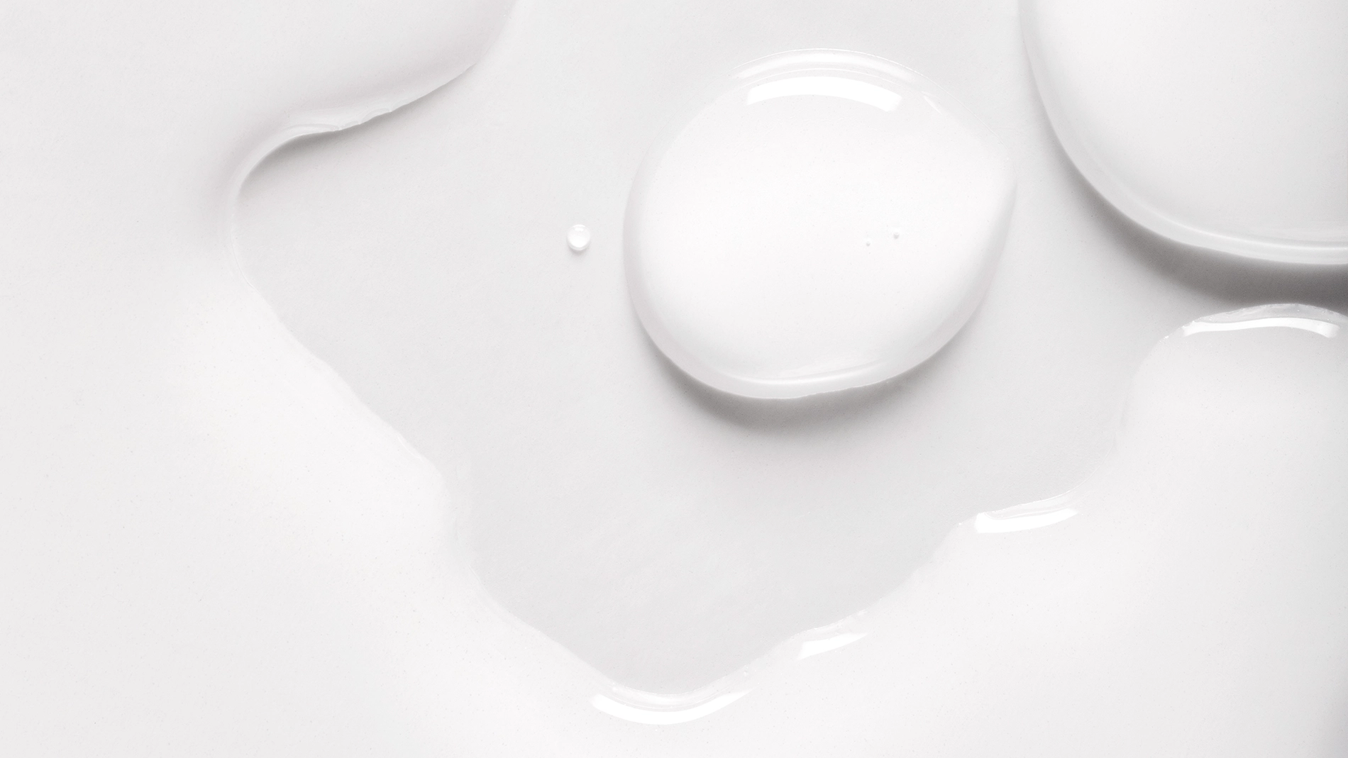 Lähikuva valkoisesta ihonhoitotuotteesta, jossa näkyy tippoja ja pehmeä, kiiltävä seerumi- tai lotion-koostumus valkoista taustaa vasten. Voidemainen rakenne korostaa kosteuttavaa skincare-tuotetta, jossa on kevyt ja silkkinen lopputulos.