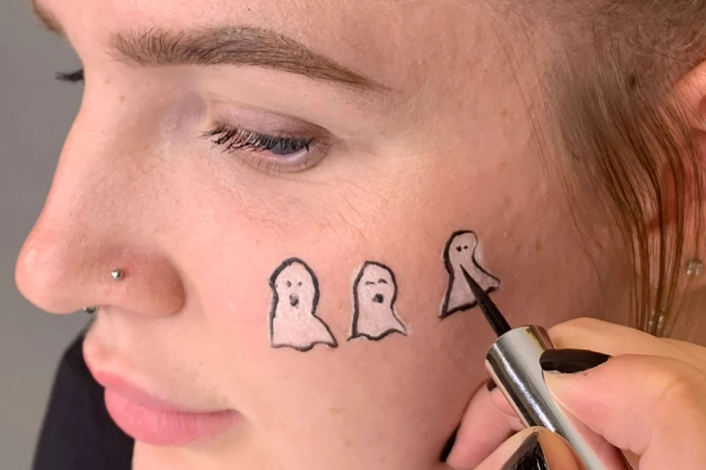 Person som tegner et ansikt på en hvit spøkelsesform på kinnet med svart flytende eyelinerpenn.