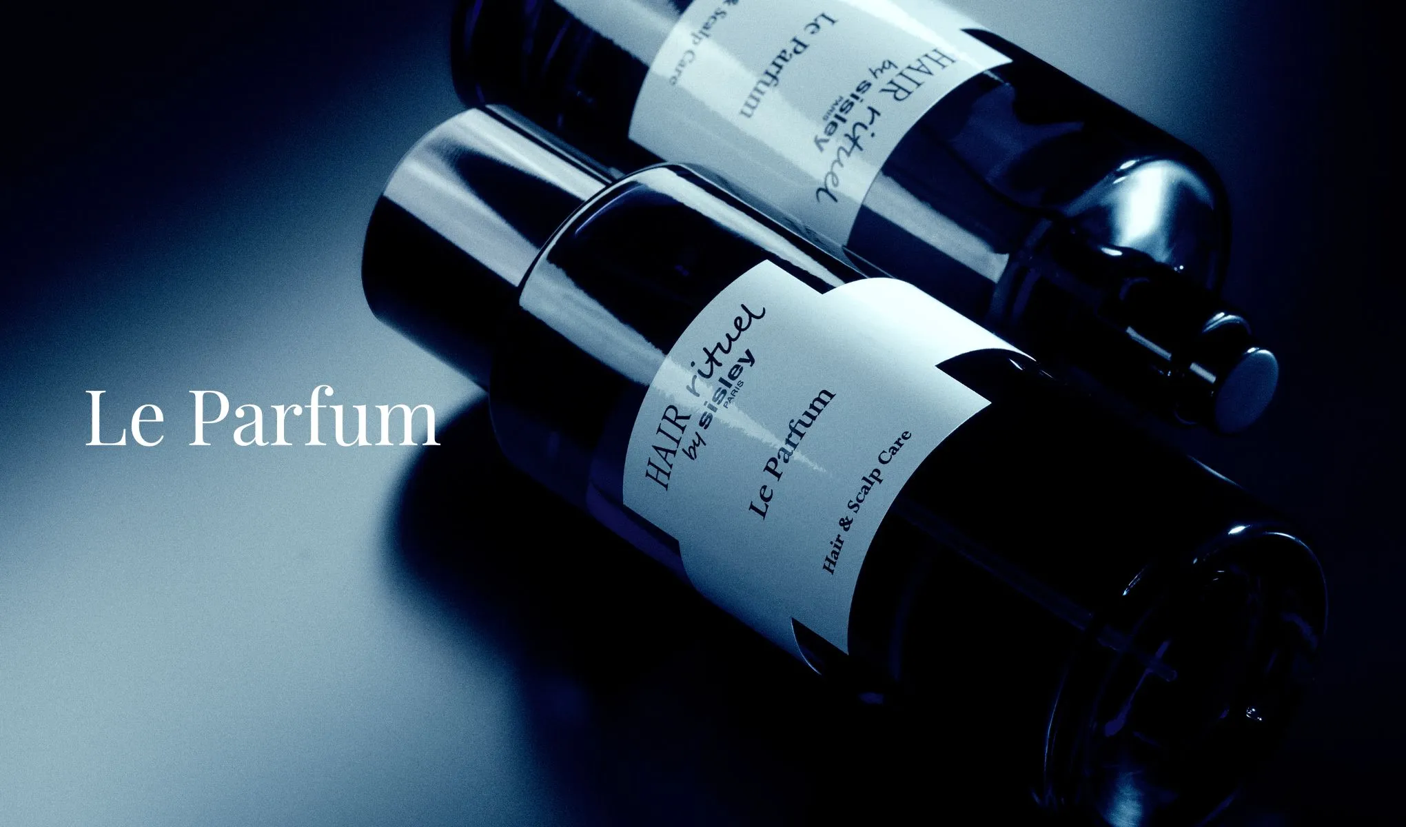 Kaksi tummaa pulloa, joissa lukee "HAIR rituel by Sisley Paris Le Parfum Hair & Scalp Care". Taustalla sinertävä valaistus ja teksti "Le Parfum" vasemmalla.