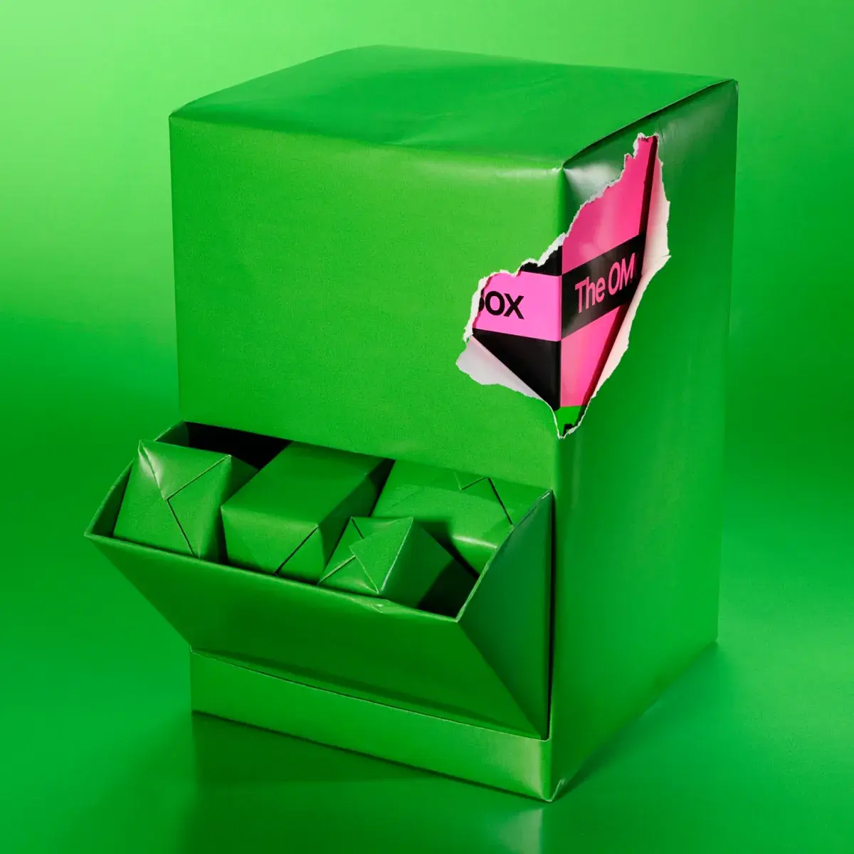 En låda inslagen i grönt papper mot en grön bakgrund. I ett öppet fack ligger små gröna paket, och en rosa och svart förpackning skymtar genom en riven öppning.