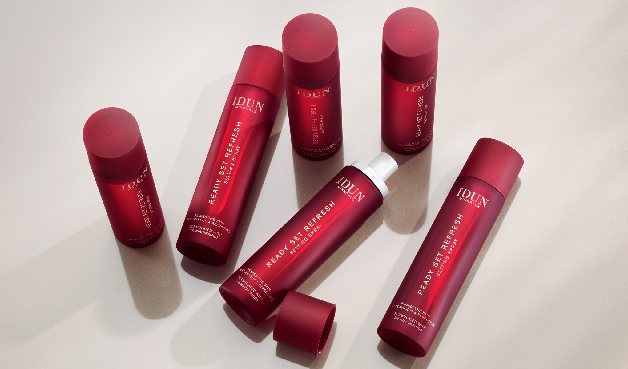 Useita punaisia pulloja IDUN Mineralsin ”Ready Set Refresh Setting Spray” -suihketta kirkkaalle pinnalle aseteltuna. Tuotteet on suunniteltu meikin virkistämiseen ja kiinteyttämiseen.