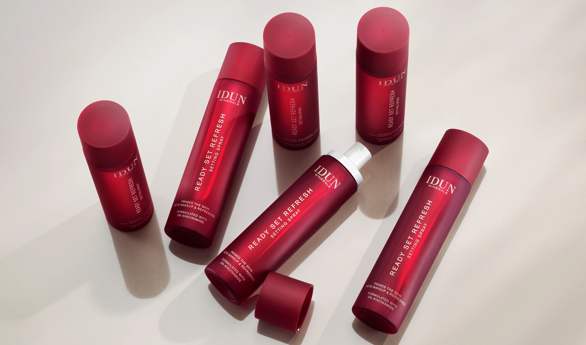 Useita punaisia pulloja IDUN Mineralsin ”Ready Set Refresh Setting Spray” -suihketta kirkkaalle pinnalle aseteltuna. Tuotteet on suunniteltu meikin virkistämiseen ja kiinteyttämiseen.