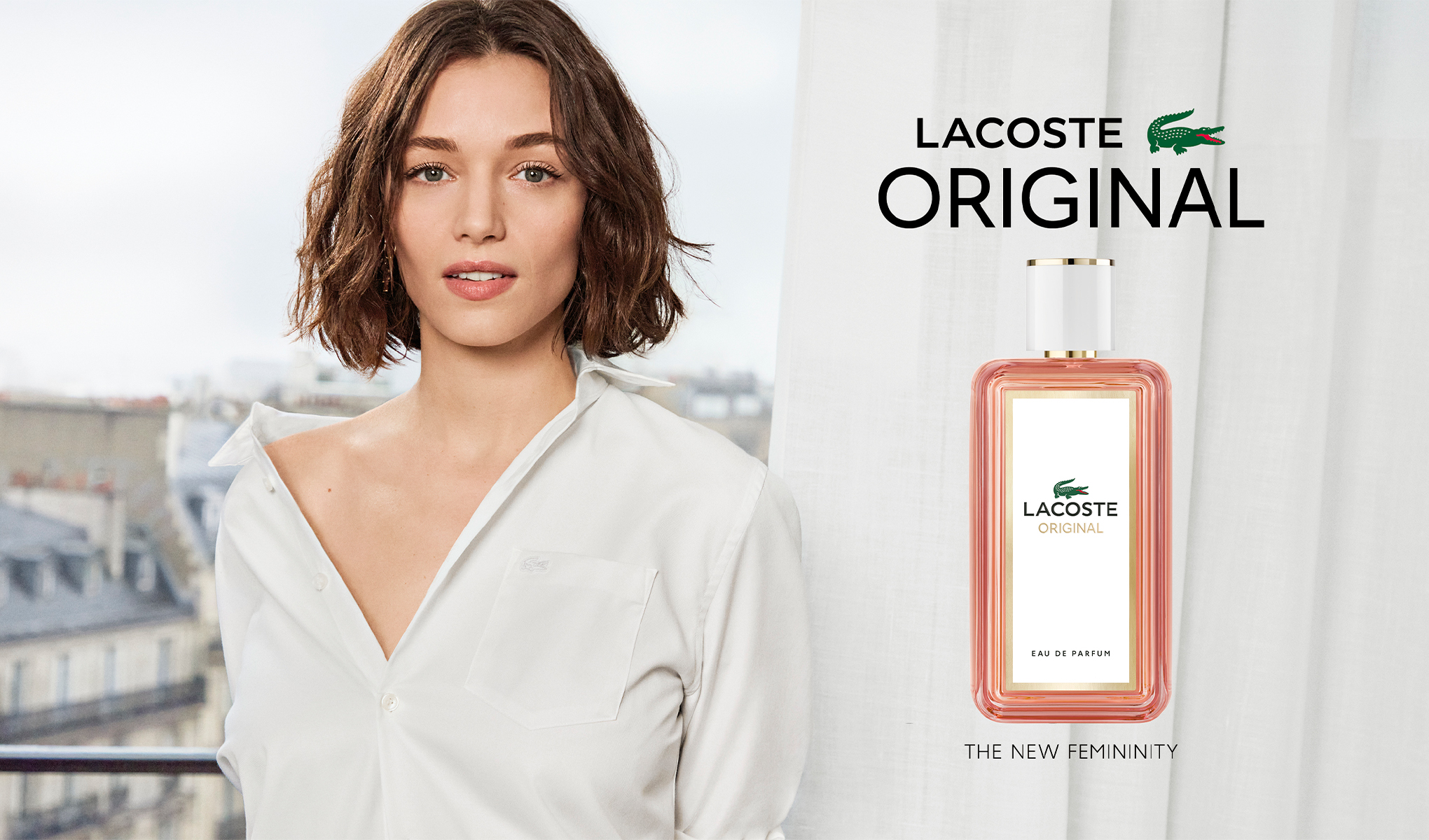 Nainen, jolla on lyhyet ruskeat hiukset ja valkoinen paita, seisoo kaupungin taustalla. Oikealla puolella on Lacoste Original -parfyymipullo ja teksti "THE NEW FEMININITY".