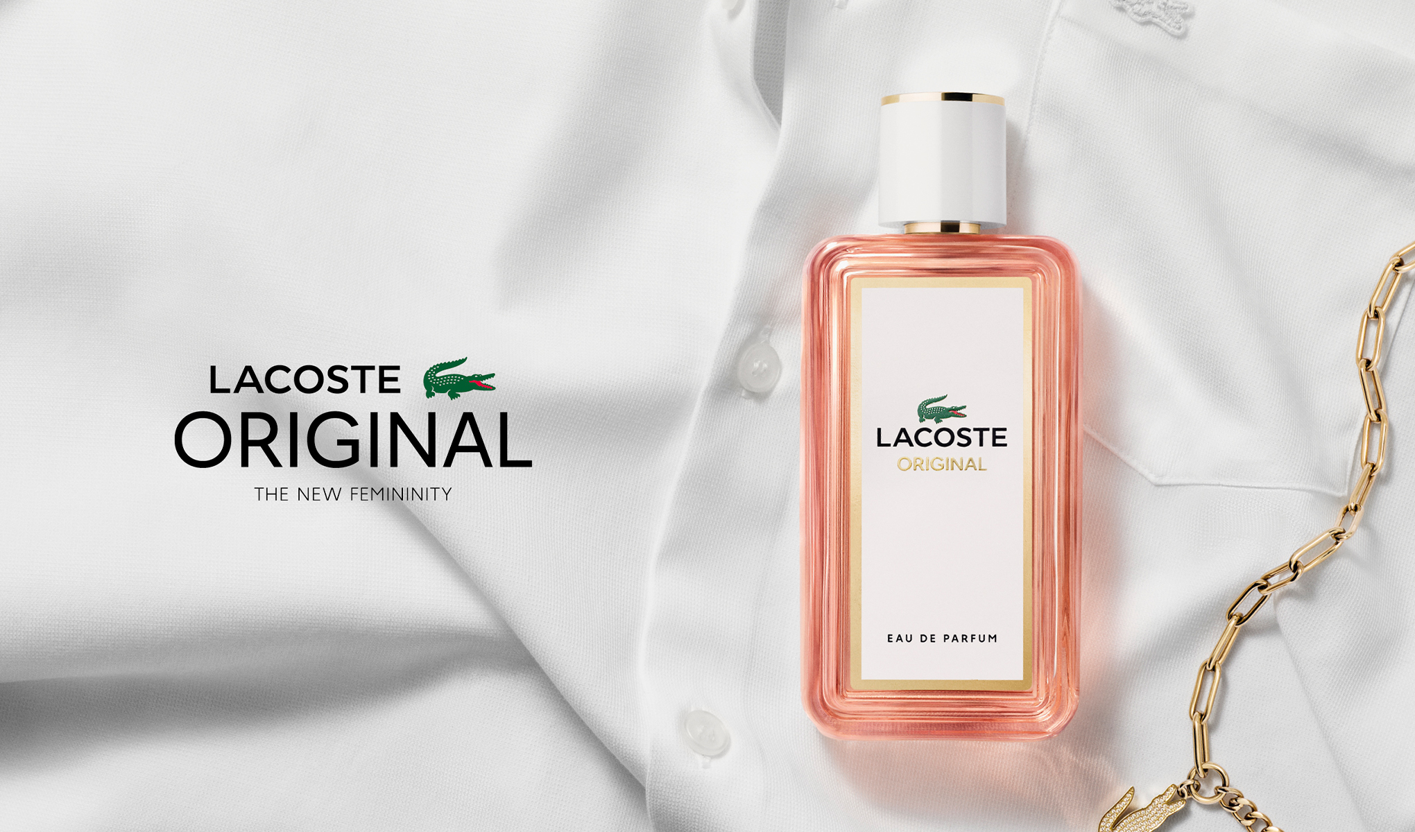 En rosa parfymeflaske fra Lacoste ligger på en hvit skjorte sammen med en gullkjede. Teksten "LACOSTE ORIGINAL" og "THE NEW FEMININITY" er synlig på venstre side av bildet.