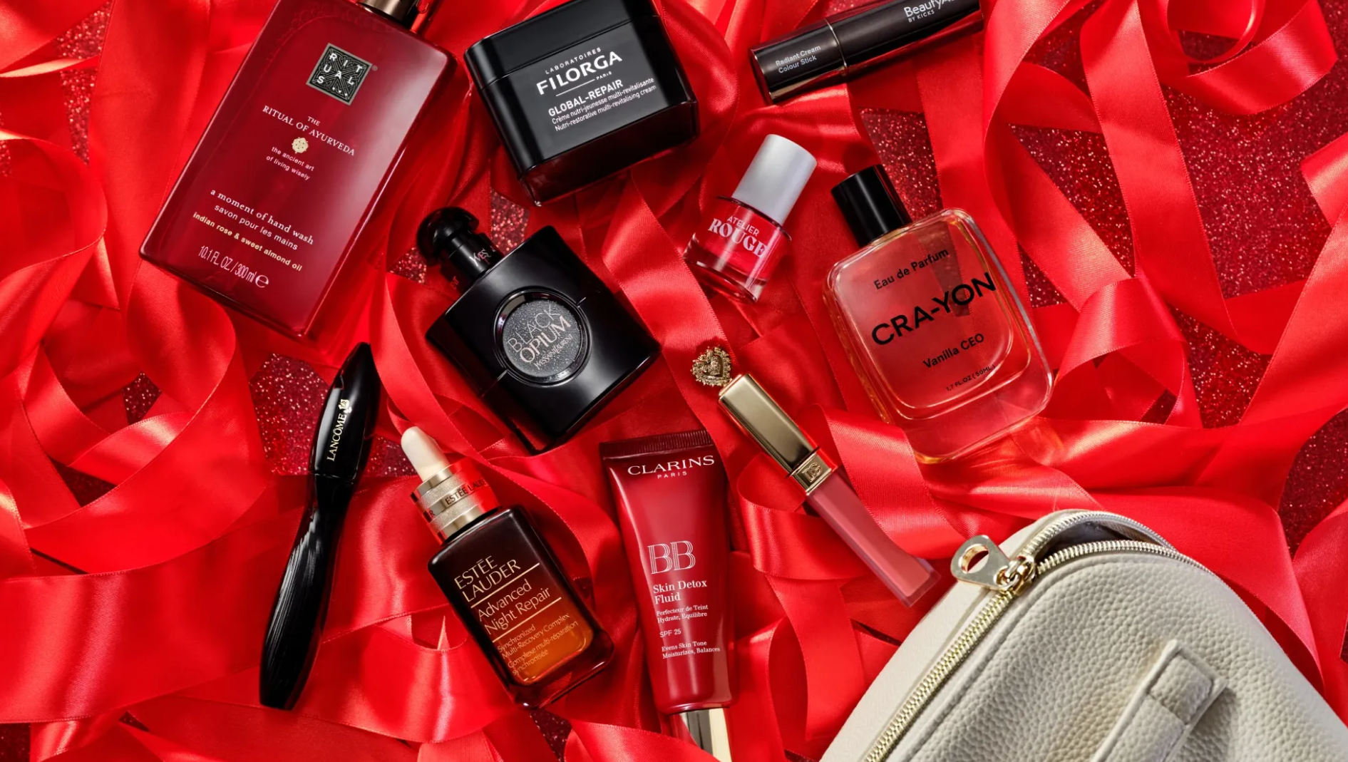 Skönhetsprodukter ligger bland röda satinband, bland annat Rituals The Ritual of Ayurveda hand wash, Filorga Global-Repair, BeautyAct Radiant Cream Colour Stick, Atelier Rouge, YSL Black Opium, CRA-YON Vanilla CEO Eau de Parfum, Lancôme mascara, Estée Lauder Advanced Night Repair, Clarins BB Skin Detox Fluid SPF 25 och en rosa läpprodukt.