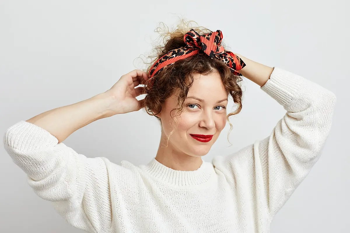 Person med krøllete hår og rød leppestift justerer et mønstret hårbånd i røde og svarte toner. Hvit strikkegenser og naturlig makeup mot lys grå bakgrunn gir et moderne beauty- og hårstylinguttrykk.