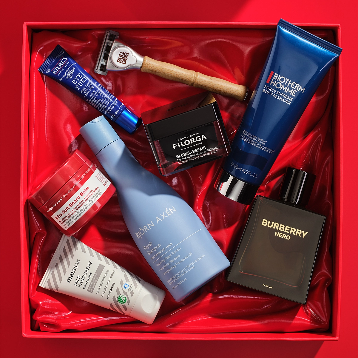 Kuva, joka näyttää useita kauneustuotteita järjestettynä punaiseen lahjapakkaukseen. Tuotteisiin kuuluu sininen tuubi Kiehl's Eye Fuel, Bulldog partahöylä, musta purkki Filorga Global-Repair, sininen tuubi Biotherm Homme Force Supreme Body Reshaper, musta pullo Burberry Hero hajuvettä, sininen pullo Björn Axén Repair Shampoo, valkoinen tuubi Matas Men kasvovoide, ja punainen purkki Recipe for Men Ultra Soft Beard Balm. Tausta on punainen.