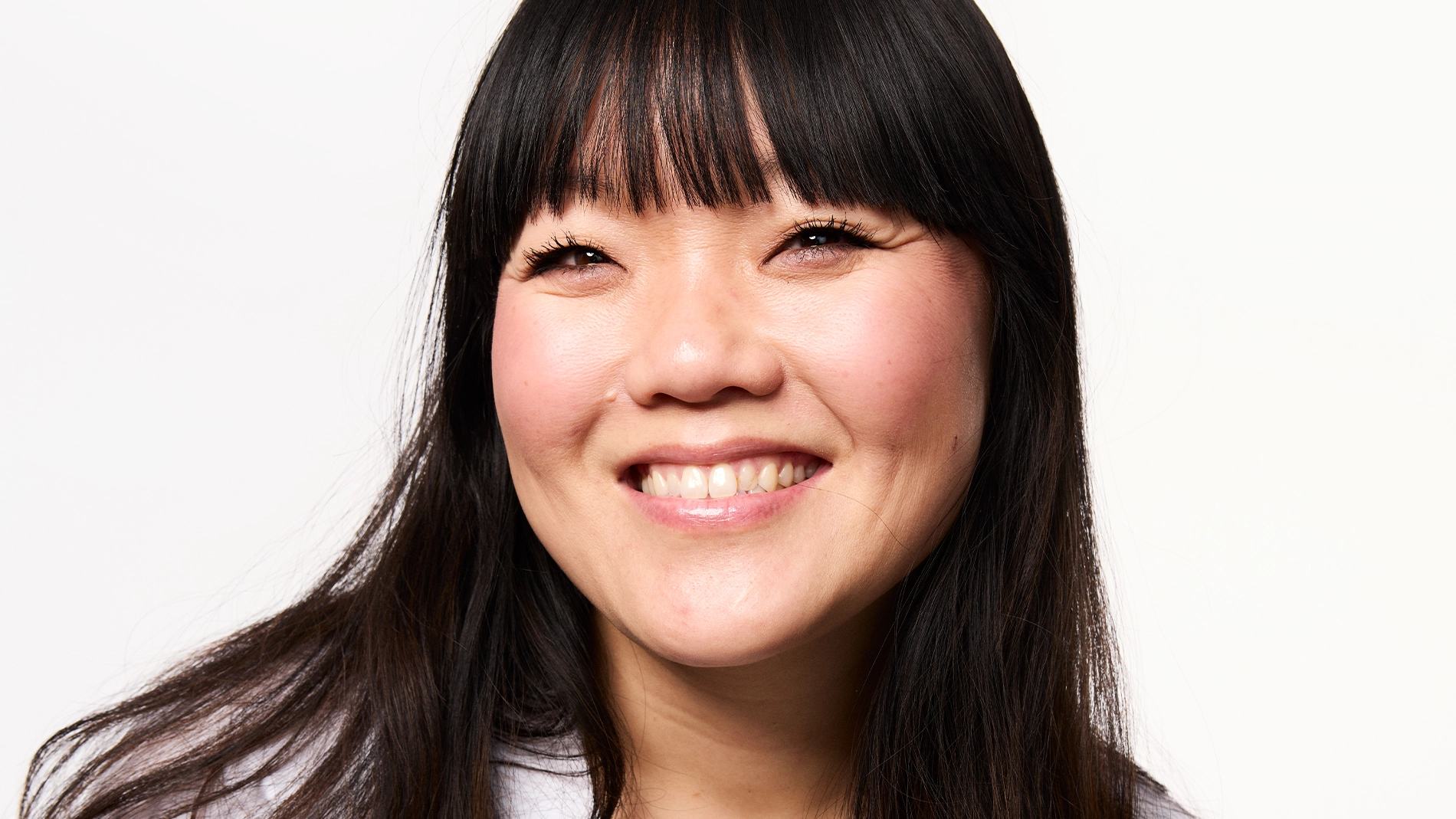 Portrett av en smilende person med langt mørkt hår og rett lugg, fotografert mot lys bakgrunn.